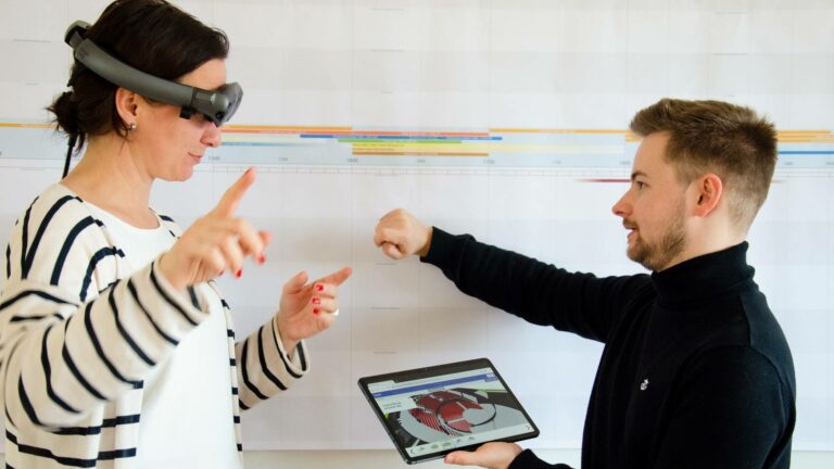 Zwei Personen, ausgestattet mit einer Mixed Reality-Brille und einem Tablet, stehen sich gegenüber. Die linke Person - eine Frau - trägt die Mixed-Reality-Brille und hebt beide Hände. Die rechte Person, ein Mann, hält mit der linken Hand ein Tablet hoch, das die 3D-Visualisierung eines Theatersaals zeigt.