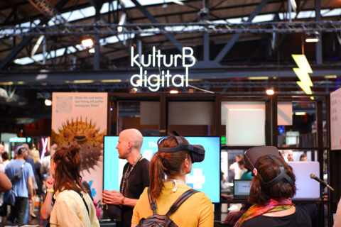 Zu sehen ist der kulturBdigital Stand auf der republica 2023. Zwei Frauen tragen VR-Brillen und sind von hinten bzw. von der Seite zu sehen. Im Hintergrund sind verschwommen weitere Personen zu sehen. In der oberen Mitte des Fotos leuchtet ein LED-Schild mit dem Schriftzug "kulturBdigital".