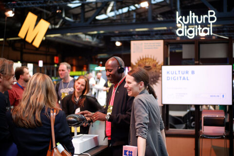 Zu sehen ist der kulturBdigital-Stand auf der republica 2023. Rechts im Bild ist ein Monitor und darüber ein LED-Schild von kulturBdigital abgebildet. Links stehen neben dem Kultursenator Joe Chialo sechs weitere Personen, teilweise mit dem Rücken zur Kamera, um ein Podest herum. Auf dem Podest steht ein taktiles Modell eines Waldmistkäfers vom Naturkunde Museum Berlin. Der Kultursenator berührt das Tastmodell und lacht dabei. Die anderen Menschen lächeln ebenfalls.