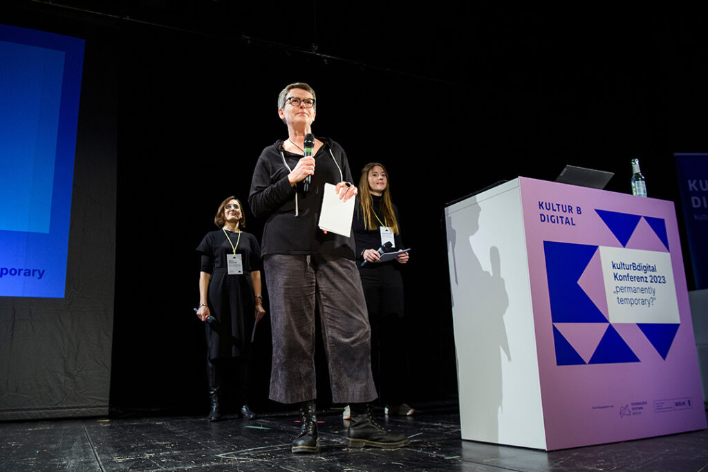 Auf einer Bühne stehen drei Frauen (Annette Kleffel, Silvia Faulstich und Lara Schulte) mit Mikrofonen in der Hand neben einem Rednerpult. Auf dem Schild am Rednerpult steht kulturBdigital Konferenz 2023, permanently temporary?