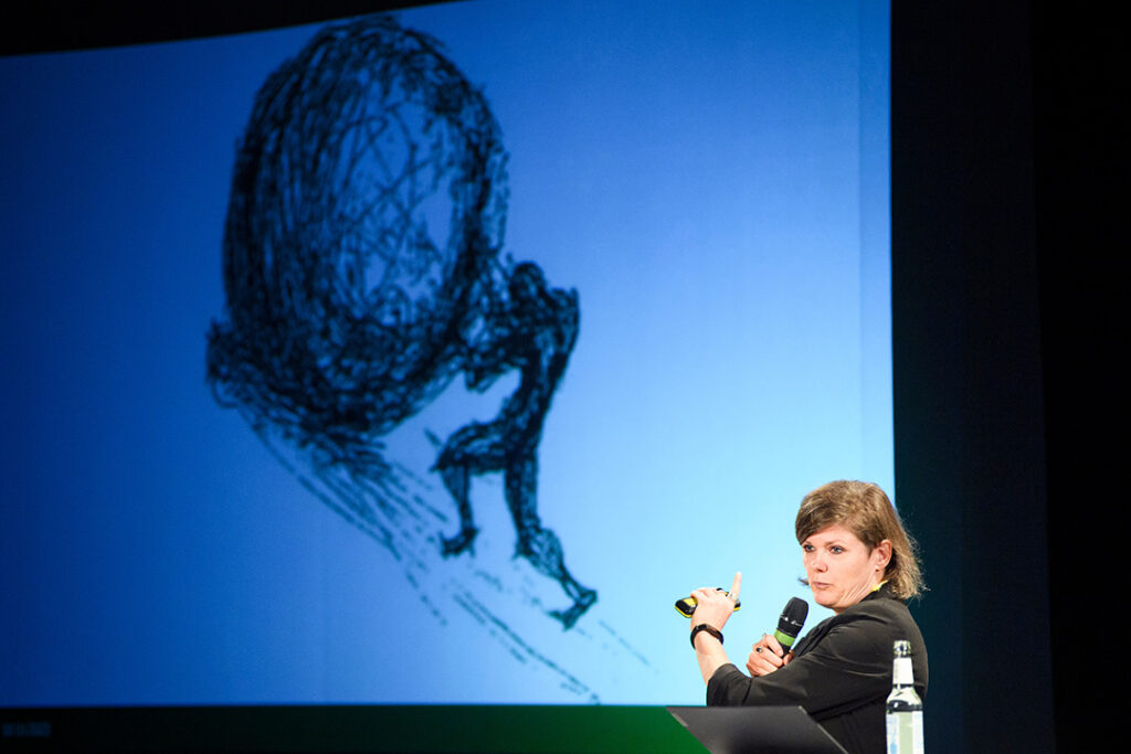 Rednerin Tina Lorenz deutet mit erhobenem Mikrofon auf eine Leinwand, auf der eine Zeichnung von Sisyphos zu sehen ist, der eine Steinkugel einen Hang hochrollt.