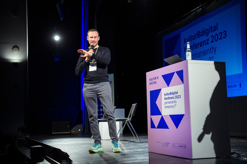 Ein Mann (Nicolas Zimmer) steht auf einer Bühne und spricht in ein Mikrofon. Auf dem Schild neben ihm steht kulturBdigital Konferenz 2023, "permanently temporary?"