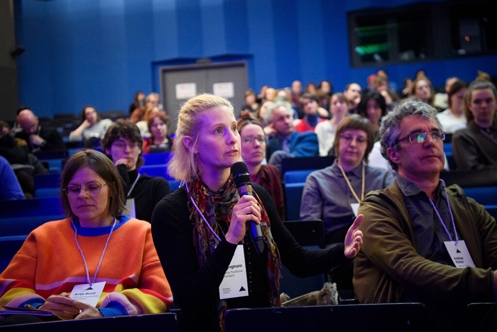 Blick in das Publikum, das in einem Theatersaal auf blauen Sitzen sitzt. Eine junge Frau mit blonden Haaren aus dem Publikum spricht in ein Mikrofon.