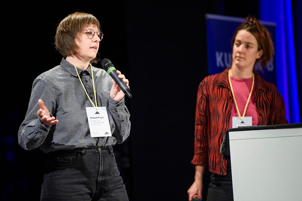 Zwei junge Frauen (Almut Pape und Leonie Rodrian) stehen mit Mikrofonen auf einer Bühne und sprechen zum Publikum.