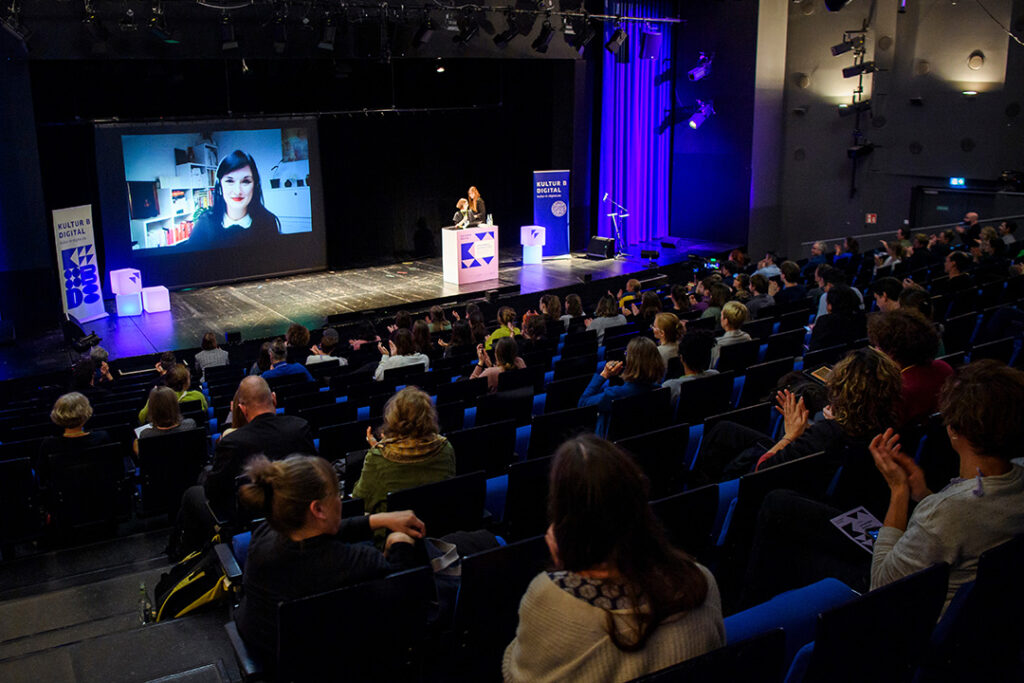 Man sieht das Publikum von hinten in Theaterreihen sitzen. Der Blick geht auf die Bühne, auf der zwei Frauen hinter einem Rednerpult stehen. In der Mitte der Bühne ist die Videoübertragung einer Frau (Tabea Schwarze) zu sehen.