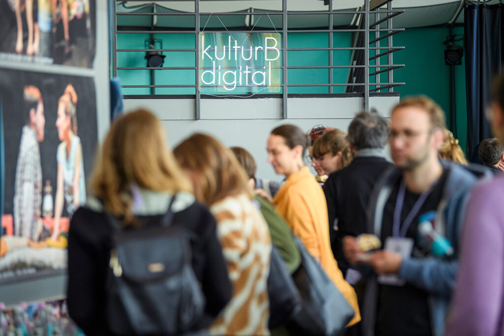 Verschwommen zu sehen ist eine Gruppe Menschen, die vor einem Leuchtschild mit den Worten kulturBdigital steht. Im Hintergrund sieht man ein Treppengeländer.