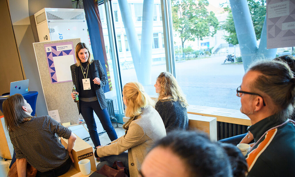 Eine Gruppe Menschen sitzt auf Hockern. Eine Frau (Julia Zimmermann) steht neben einer Pinnwand und spricht.