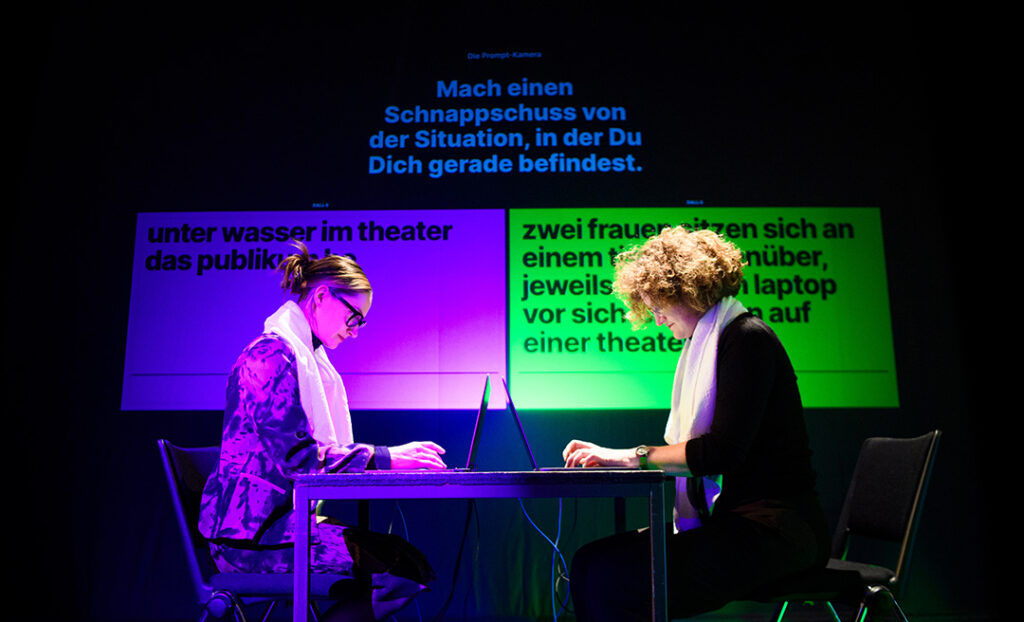 Zwei Frauen sitzen auf einer Bühne, jeweils in die Tastatur eines Laptops tippend. Hinter ihnen ist eine Aufgabe eingeblendet sowie die jeweiligen Lösungen, an denen die Frauen arbeiten.