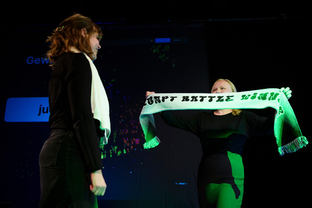 Zu sehen sind zwei junge Frauen. Die rechte Frau hält einen Schal mit der Aufschrift "Prompt Battle Winner" hoch und überreicht ihn der linken Frau.
