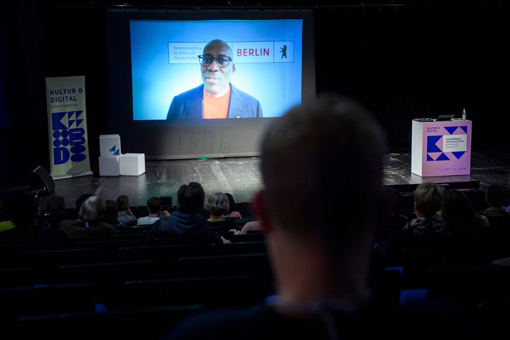 Auf einer Leinwand auf der Bühne des ATZE Musiktheaters ist Kultursenator Joe Chialo zu sehen. Das projizierte Video zeigt ihn frontal in die Kamera blickend. Im Vordergrund sind Teile des Publikums in Rückansicht zu erkennen.