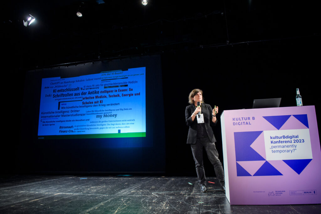 Rednerin Tina Lorenz auf der Bühne, hinter ihr eine Leinwand, auf der eine Collage aus verschiedenen Schlagzeilen zum Thema Künstliche Intelligenz zu sehen ist.
