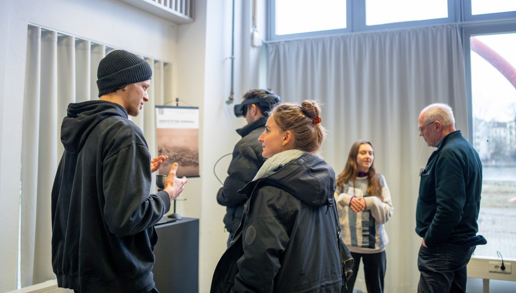 Ein junger Mann steht vor der Präsentation seines XR-Projekts. Er spricht mit einer jungen Frau und gestikuliert, sie hört ihm zu. Im Hintergrund stehen weitere Personen in einer kleinen Gruppe zusammen, ein Mann trägt eine VR-Brille.