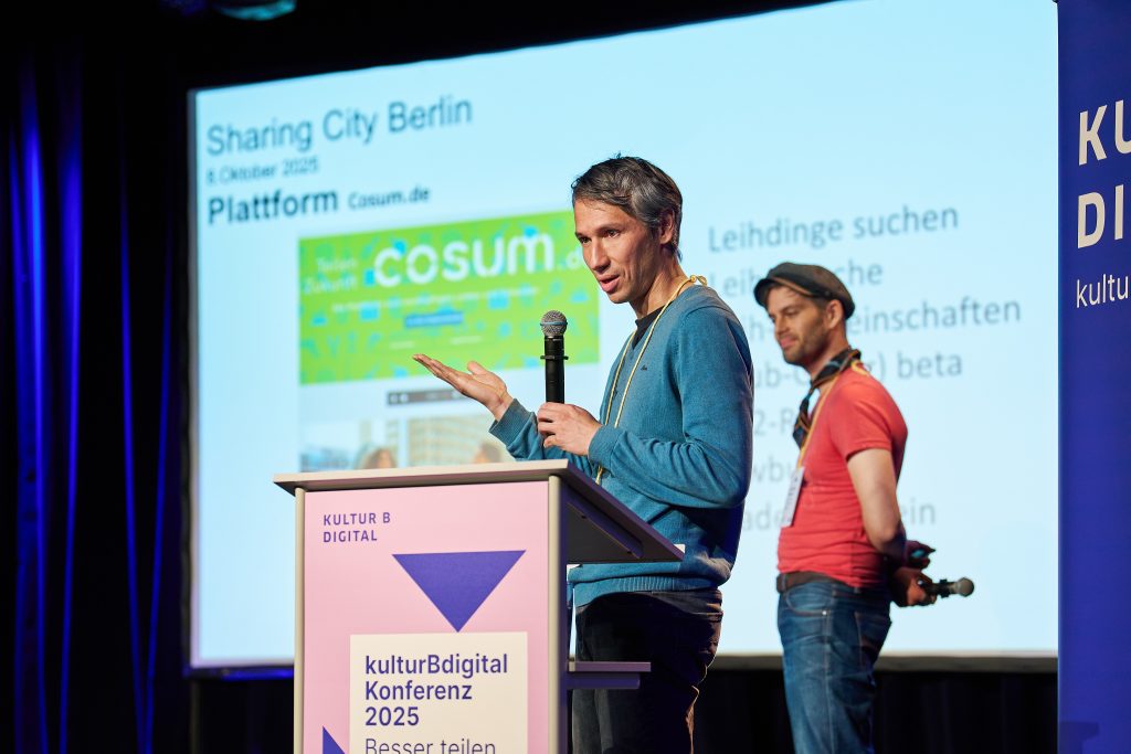 Nikolai Wolfert steht am Rednerpult auf der Bühne der kulturBdigital-Konferenz, gestikuliert und spricht ins Mikro. Jan Philip Steimel steht lächelnd rechts hinter ihm. Hinter den beiden eine Folie mit der Überschrift: Sharing City Berlin.