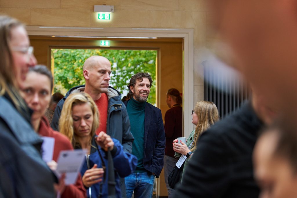 Mehrere Teilnehmende stehen in der Einlassschlange, einige unterhalten sich. Im Hintergrund ein geöffneter Ausgang mit Blick ins Grüne.