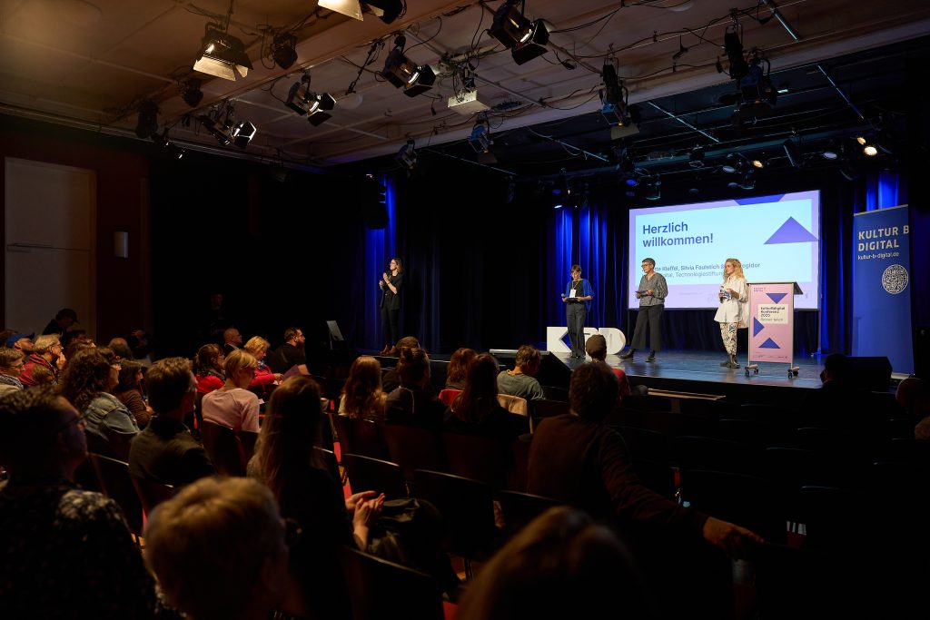 Silvia Faulstich, Annette Kleffel, Ana Regidor blicken von der Bühne ins Publikum, hinter ihnen eine Leinwand mit der Aufschrift ‚Herzlich willkommen!‘. Am linken Bühnenrand ist die Gebärdensprachdolmetscherin bei ihrer Arbeit zu sehen.