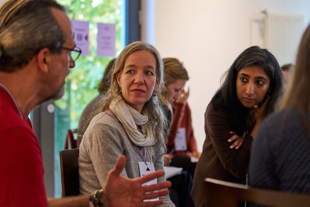 Mehrere Personen sitzen in einer Gesprächsrunde in einem hellen Raum, eine Person gestikuliert mit ausgestreckter Hand.