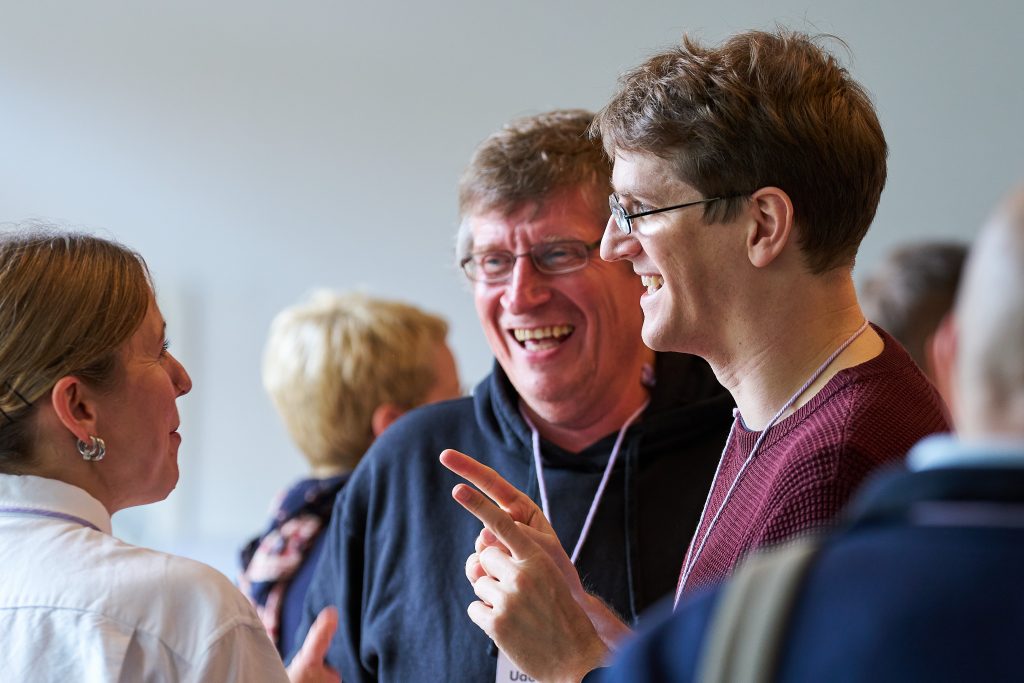 Drei Teilnehmende stehen im Foyer, schauen belustigt und lachen einander an. Eine Person gestikuliert.
