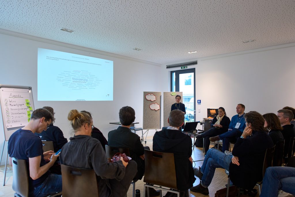 Teilnehmende sitzen im Stuhlkreis in einem Seminarraum und blicken auf eine Präsentation mit einer Wortwolke. Im Mittelpunkt der Wortwolke steht Finanzierung und Wissenstransfer.
