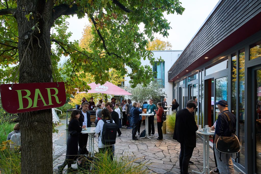 Menschen stehen im Garten vor einem Veranstaltungsgebäude an Stehtischen, ein rotes Schild mit der Aufschrift ‚Bar‘ hängt an einem Baum.