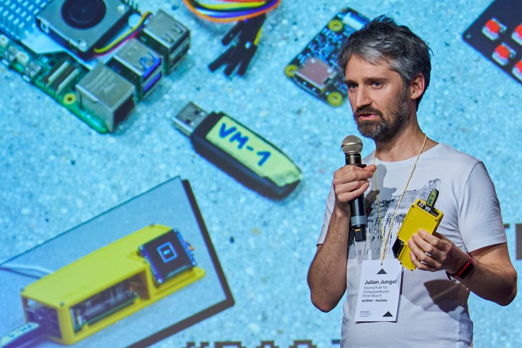 Julian Jungel hält ein gelbes Elektronikgerät in der Hand und spricht ins Mikrofon, im Hintergrund sind verschiedene Hardware-Komponenten auf der Folie einer Präsentation zu sehen.