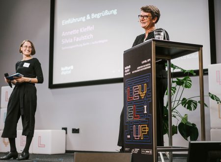 Silvia Faulstich (links) und Annette Kleffel (rechts) vom Team kulturBdigital stehen auf einer Bühne während der kulturBdigital Konferenz 2022. Annette spricht die Grußworte, wie man der Projektionsfläche im Hintergrund an entnehmen kann. Sie blickt in Richtung Publikum.