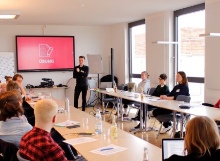 Video-Workshop bei kulturBdigital: In einem Workshopraum der Technologiestiftung Berlin sitzen 13 Personen an einem Tusch-U verteilt. Ein Redner befindet sich in der Mitte vor einem Videobildschirm. Darauf steht das Wort Übung.